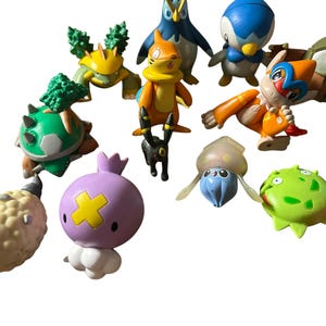 May include: A collection of small plastic character figurines featuring various creatures in colors including orange, green, blue, purple, and beige. The figures display diverse shapes, some with spiky textures, smooth surfaces, and distinct facial features.