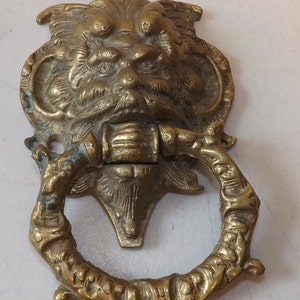 May include: Antique brass door knocker featuring a detailed face with a beard and a ring-shaped handle. The knocker has an aged, weathered appearance, suggesting it is vintage. The design is ornate and decorative, suitable for a front door.