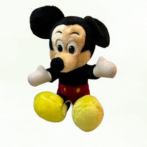 Puede incluir: Un peluche de Mickey Mouse con orejas negras, cuerpo negro y cara beige. El juguete tiene guantes blancos, pantalones cortos rojos y zapatos amarillos. El juguete es un personaje cl&aacute;sico de Disney.