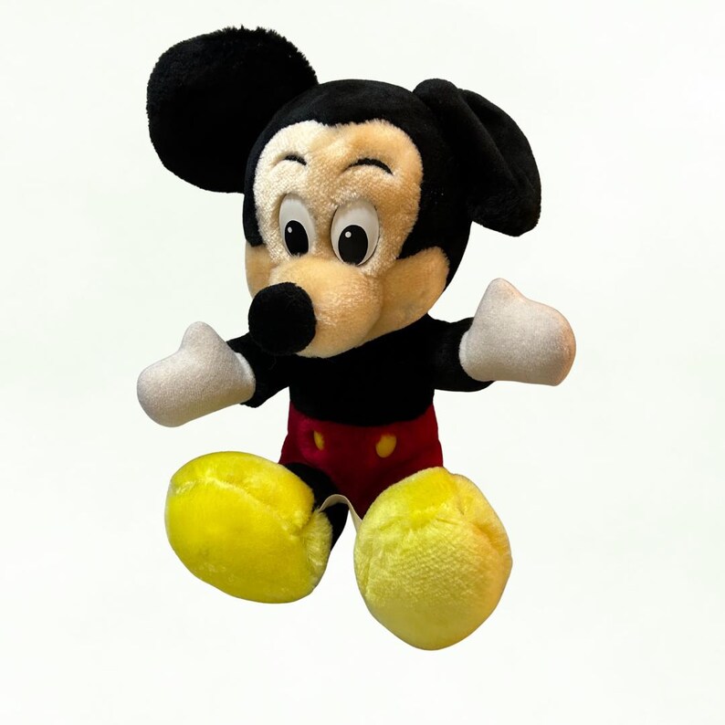 Puede incluir: Un peluche de Mickey Mouse con orejas negras, cuerpo negro y cara beige. El juguete tiene guantes blancos, pantalones cortos rojos y zapatos amarillos. El juguete es un personaje cl&aacute;sico de Disney.