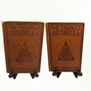 Peut inclure: Deux livres anciens intitulés "The Triangle Arithmetics" sont présentés. Les couvertures sont marron avec des lettres noires et un motif triangulaire central. Un livre est intitulé "Grade Eight", l'autre "Grade Seven".