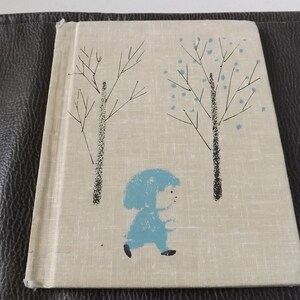 May include: A vintage book with a light beige cover featuring a whimsical illustration. The cover art depicts a child in a blue coat walking between two stylized trees with black trunks and sparse branches. The background is a textured, off-white.