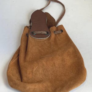 May include: A small, brown suede pouch with a drawstring closure. The pouch has a leather flap and a thin, brown cord. A small, silver-coloured bead is attached to the end of the cord. The pouch is designed to be carried or worn.