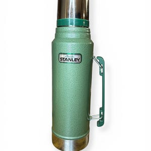 May include: A vintage green Stanley thermos with a silver lid and base. The thermos has a green handle and the "Stanley" logo is visible. The thermos is designed to keep liquids hot or cold.
