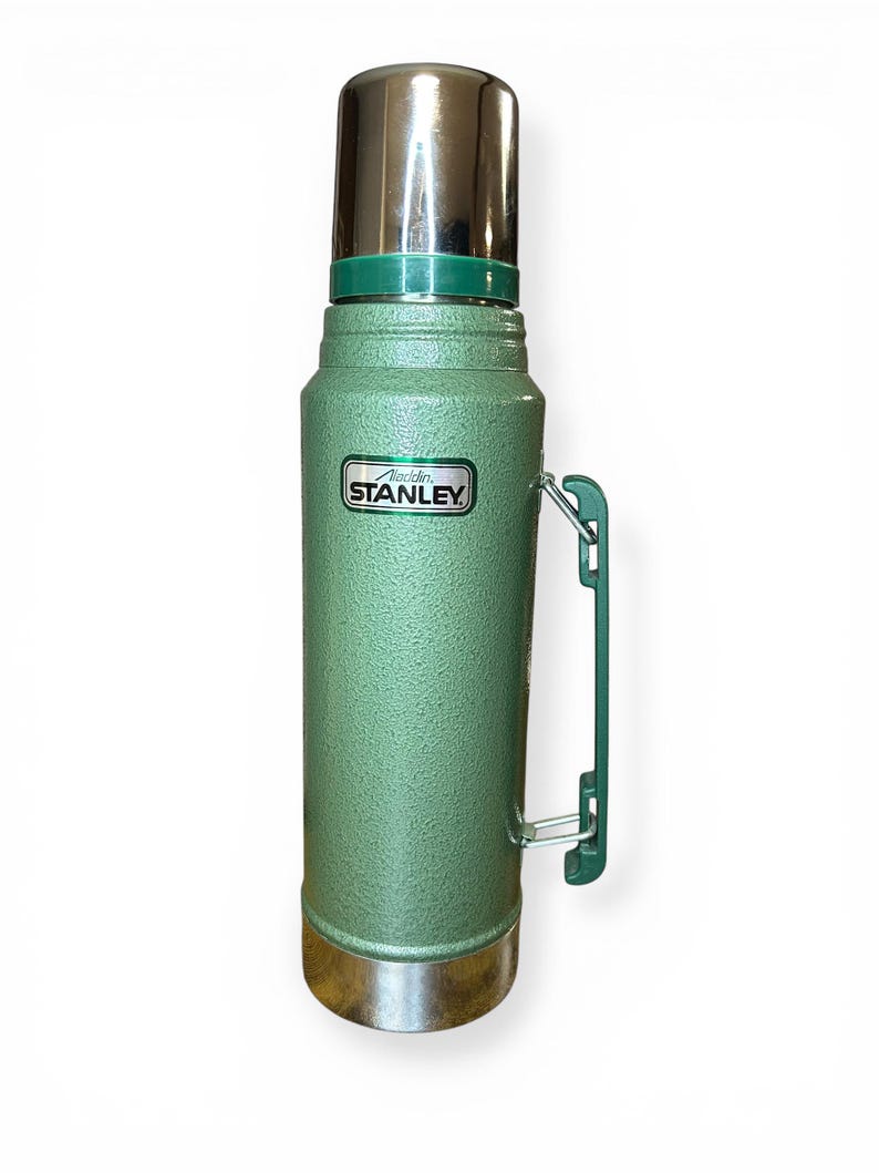 May include: A vintage green Stanley thermos with a silver lid and base. The thermos has a green handle and the "Stanley" logo is visible. The thermos is designed to keep liquids hot or cold.