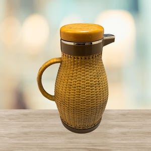 May include: A vintage insulated pitcher with a woven rattan exterior. The pitcher has a wooden lid and handle, with a brown band around the middle. The spout is silver-colored. The pitcher sits on a wooden surface.