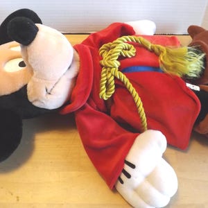 May include: A plush Mickey Mouse dressed as a sorcerer, lying down. Mickey wears a blue wizard hat with a white crescent moon, a red robe with a gold rope, and brown boots. The plush toy has black ears and a tan face.