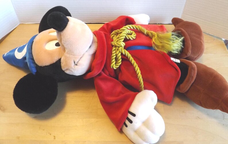 May include: A plush Mickey Mouse dressed as a sorcerer, lying down. Mickey wears a blue wizard hat with a white crescent moon, a red robe with a gold rope, and brown boots. The plush toy has black ears and a tan face.