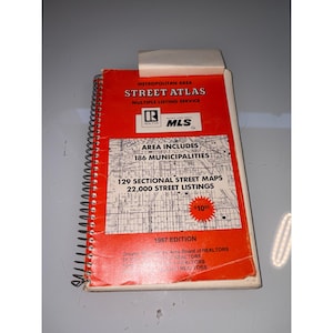 以下が含まれることがあります： Multiple Listing Service発行の1987年版メトロポリタンエリアストリートアトラス。スパイラル綴じのオレンジ色の表紙には、黒い文字、Realtorのロゴ、地図のグラフィック、そして10.95ドルの価格が印刷された赤い星型のグラフィックが描かれています。
