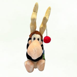 Pode incluir: Um brinquedo de pelúcia de alce com grandes chifres bege, um chapéu marrom e um nariz grande rosa claro. O alce tem um corpo preto e branco e uma pequena bola vermelha pendurada em um chifre. Uma etiqueta está presa ao alce.