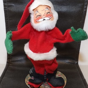 May include: A vintage Santa Claus figurine with a red suit, white faux fur trim, and a matching hat. The figure has a painted face, white beard, and green mittens. It stands on a round base with a floral pattern, and black boots.