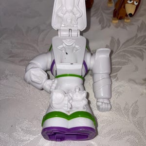 Puede incluir: Una figura de acci&oacute;n blanca de Buzz Lightyear con detalles verdes, morados y negros. La figura tiene una cabeza con bisagras y una peque&ntilde;a figura alien&iacute;gena en la parte delantera. Un juguete Slinky Dog marr&oacute;n y canela est&aacute; en el fondo.
