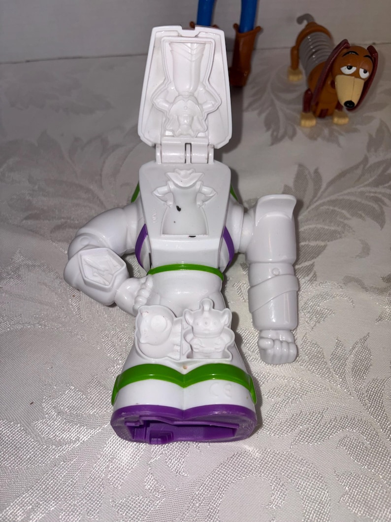Puede incluir: Una figura de acci&oacute;n blanca de Buzz Lightyear con detalles verdes, morados y negros. La figura tiene una cabeza con bisagras y una peque&ntilde;a figura alien&iacute;gena en la parte delantera. Un juguete Slinky Dog marr&oacute;n y canela est&aacute; en el fondo.