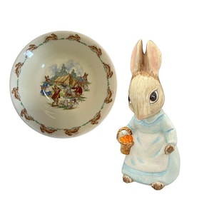 May include: A ceramic bowl with a rim decorated with brown rabbit silhouettes and a central illustration of rabbits picnicking. Beside it is a ceramic rabbit figurine holding a basket of carrots, wearing a blue dress and white apron.