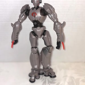 May include: A gray action figure of a robot with red accents. The robot has a Cobra symbol on its chest and is posed with its arms out. The figure has a detailed design with multiple joints and a helmet.