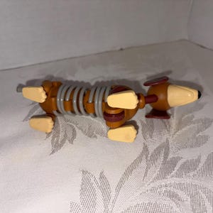 Puede incluir: Un perro salchicha de juguete con un cuerpo en forma de resorte. El perro es marr&oacute;n con patas beige y un hocico beige. El cuerpo es un resorte gris. Las orejas del perro son marrones y tiene una nariz negra.