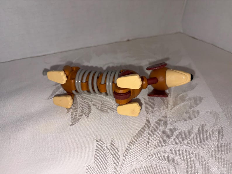 Puede incluir: Un perro salchicha de juguete con un cuerpo en forma de resorte. El perro es marr&oacute;n con patas beige y un hocico beige. El cuerpo es un resorte gris. Las orejas del perro son marrones y tiene una nariz negra.