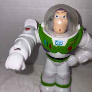 Puede incluir: Figura de acci&oacute;n de Buzz Lightyear de la franquicia Toy Story. La figura es principalmente blanca con detalles verdes y morados. Cuenta con un casco transparente, una placa pectoral verde con las palabras "Space Ranger" y "Lightyear", y brazos articulados.
