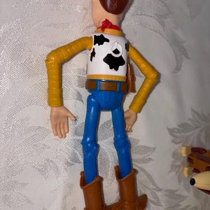 Puede incluir: Figura de acci&oacute;n de Woody, vista desde atr&aacute;s, con sombrero de vaquero marr&oacute;n, chaleco blanco con manchas negras, camisa amarilla, pantalones azules y botas de vaquero marrones. La figura tiene brazos y piernas articulados.