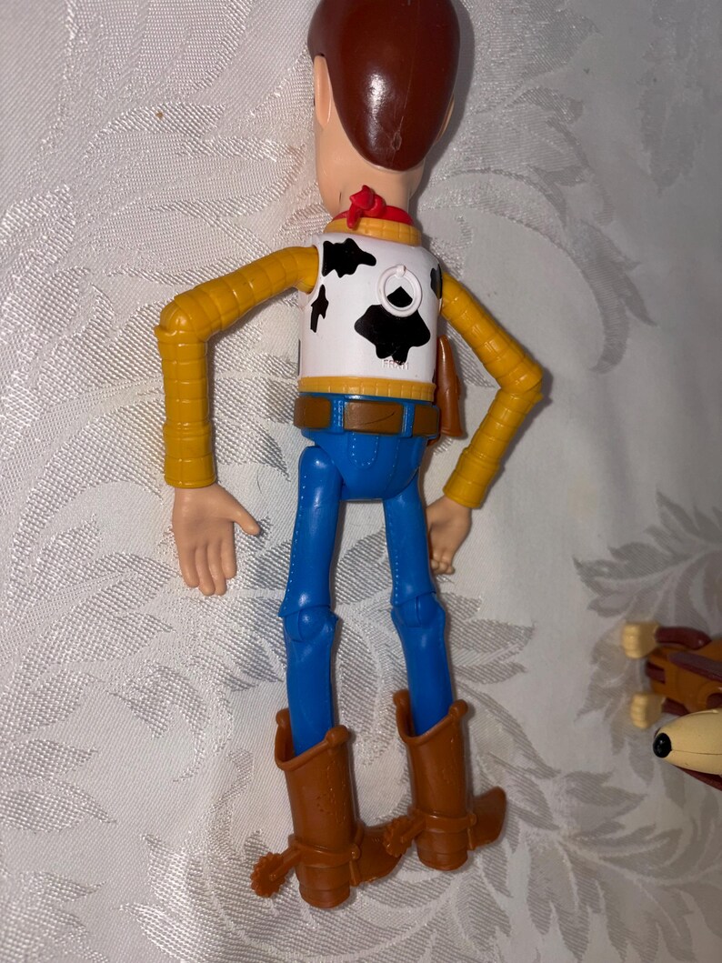 Puede incluir: Figura de acci&oacute;n de Woody, vista desde atr&aacute;s, con sombrero de vaquero marr&oacute;n, chaleco blanco con manchas negras, camisa amarilla, pantalones azules y botas de vaquero marrones. La figura tiene brazos y piernas articulados.