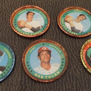 May include: A collection of vintage baseball player tokens. Each circular token features a player's portrait, team name, and position. The tokens have a metallic rim and a colorful design. Players include Gaylord Perry, Joe Torre, Bob Tolan, and Reggie Jackson.