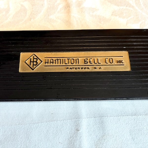 May include: A black rectangular box with a gold-colored metal plate. The plate features the text "HAMILTON BELL CO. INC." and "PATERSON, N.J." in black lettering. The box has a ribbed texture.