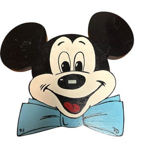 May include: A wooden wall clock shaped like Mickey Mouse's head, featuring black ears, a cream-coloured face, and a light blue bow tie. The clock has a digital display on the nose, the number 93 on the left, and the initials RD on the right.