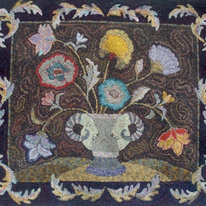 May include: A rug hooking depicting a vase with a ram's head design, filled with colorful flowers. The vase is on a brown background with a dark blue border and a floral design.