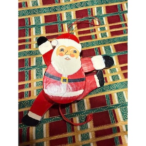 May include: A wooden Santa Claus ornament featuring a red suit with a black belt and gold buckle, white beard, and black boots. The figure is in a dynamic pose with one arm and one leg extended, and includes a red string for hanging and a pull cord.