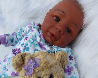 May include: A realistic baby doll with dark skin, dressed in a floral onesie in white, teal, and purple. Beside the doll is a beige teddy bear decorated with purple flowers and a pink ribbon.