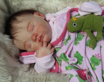 May include: A realistic baby doll with closed eyes, wearing a pink and white striped outfit and a matching robe. A green frog plush toy with a crown rests on the blanket. The doll is lying on a fluffy, white surface.
