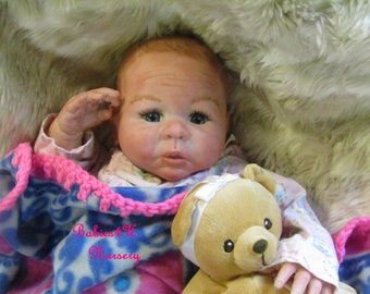 May include: A realistic baby doll with fair skin, blue eyes, and light brown hair, resting on a soft blanket. The doll is holding a small teddy bear with a bandage on its head. The blanket has a pink and blue pattern, and the text "Babies 4 U Nursery" is visible.