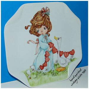 May include: Hand-painted porcelain plate featuring a girl with brown hair and a blue dress holding a string of red hearts. A white goose with yellow feet is walking beside her. The plate has the text "hand painted vintage plate 1992" on the bottom right corner.