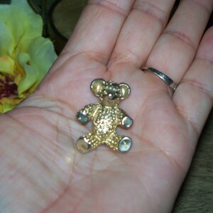 May include: A gold-colored teddy bear pin with a textured surface. The pin is sitting on a person's hand.