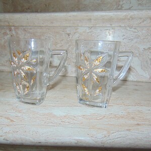 May include: Two clear glass mugs with gold floral designs. The mugs have a square shape and a handle on the side.