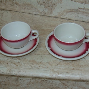 May include: Two white ceramic teacups with red trim and matching saucers. The cups and saucers have a red band around the rim.