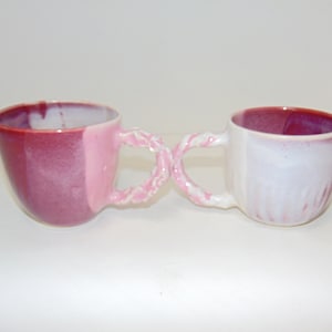 May include: Two ceramic mugs with pink and white glaze, connected by a single, intertwined handle. The mugs are a light pink and white color.