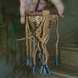May include: A small, woven basket with leather straps and metal bells hanging from the bottom. The basket is made of natural materials and has a rustic, handmade look.
