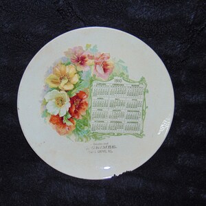 May include: A white ceramic plate with a floral design and a calendar for the year 1910. The calendar is surrounded by a green vine border. The plate is decorated with a floral design of yellow, white, and red flowers. The text "Souvenir of G. Grimes, 541 S. Grove, Ill." is printed on the bottom of the plate.