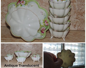 May include: Antique translucent Nippon serving set. The set includes a large, scalloped serving dish and six small footed bowls. The white porcelain pieces have a green and gold trim with pink floral accents. The text "Antique Translucent Nippon" is visible.