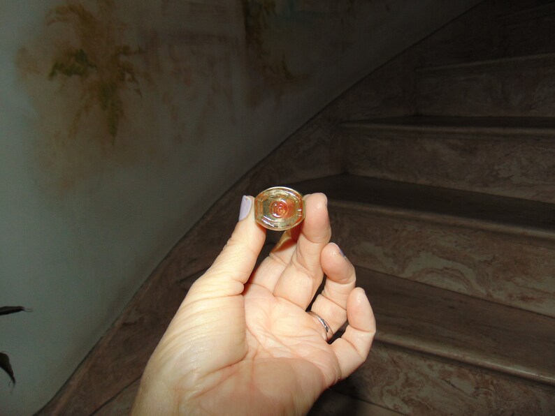 May include: A close-up of a gold-colored bottle stopper with a clear, round top. The stopper has a red, circular design in the center.