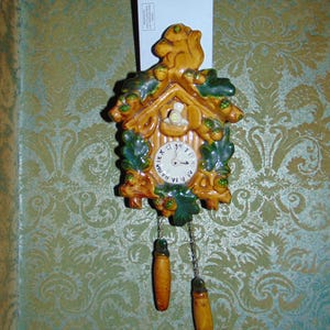 May include: A ceramic cuckoo clock with a squirrel on top and acorns decorating the sides. The clock face has Roman numerals and a pendulum with two wooden weights.