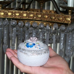 May include: A white porcelain trinket box with a floral design. The box has a raised, textured surface and a lid with a floral design in shades of blue, pink, and green.