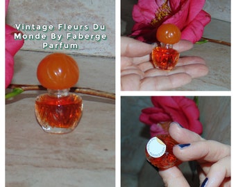 May include: Vintage miniature bottle of Fleurs Du Monde By Faberge Parfum. The bottle is clear glass with a textured design and an orange glass stopper. The bottle is sitting on a light brown surface.