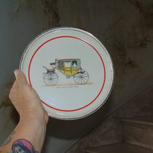 May include: A white ceramic plate with a red rim and a gold trim. The plate features a black and white illustration of a horse-drawn carriage with yellow accents.