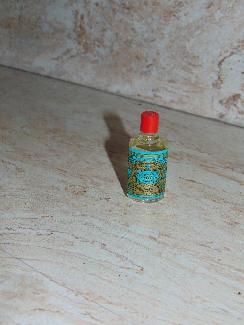 May include: A small, clear glass bottle with a red cap and a blue and gold label. The label reads "Eau de Cologne" and "4711" in a decorative font.