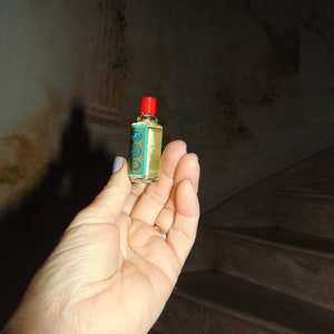 May include: A small, clear glass bottle with a red cap and a blue and gold label. The bottle is filled with a yellow liquid.