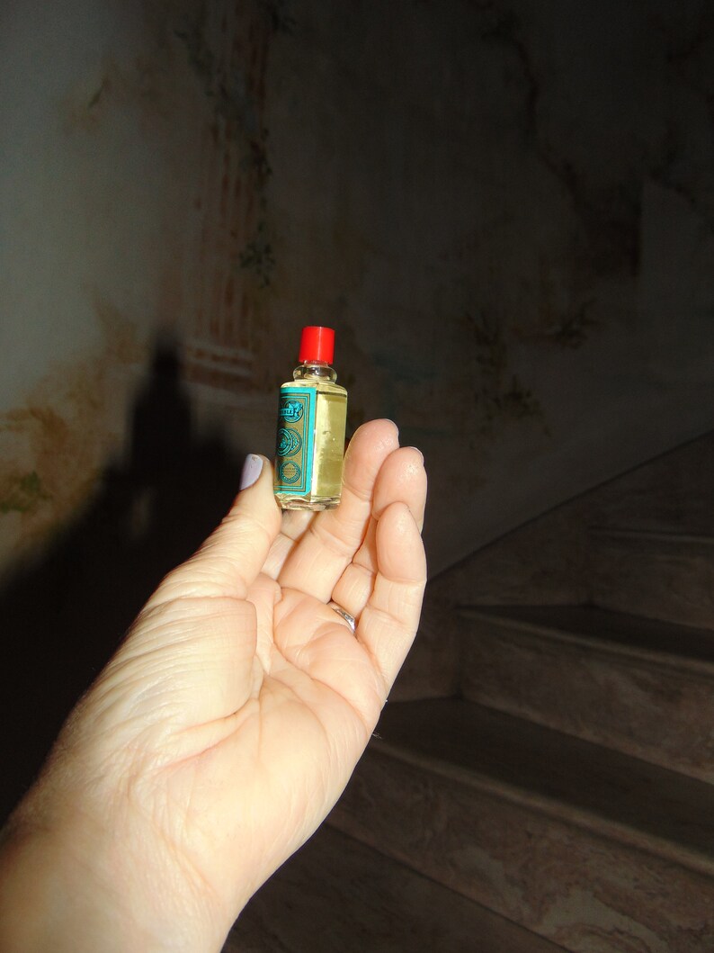 May include: A small, clear glass bottle with a red cap and a blue and gold label. The bottle is filled with a yellow liquid.