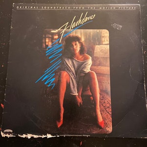 May include: A black vinyl record album cover with the title "Flashdance" in blue and white lettering. The cover features a woman in a grey sweater and red shoes sitting on a chair. The album is the original soundtrack from the motion picture.