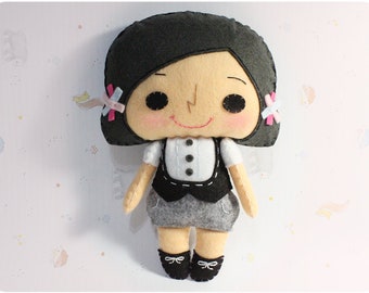 May include: A handmade felt doll with black hair, a white shirt, a black vest, and a gray skirt. The doll has a smiling face with black eyes and pink cheeks.